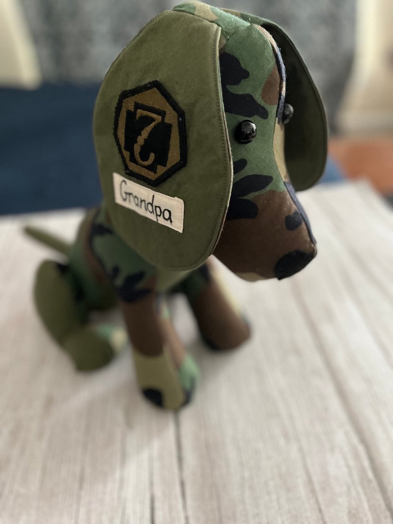 A keepsake military dog made from an upcycled army coat is sitting on a bed.  The dog has an army medic badge sewn on one ear.