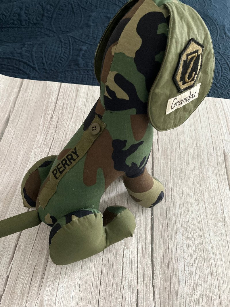 A keepsake military dog made from an upcycled army coat is sitting on a bed.  The dog has an army medic badge sewn on one ear.